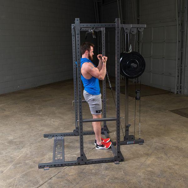 Four-post Powerline Half Rack PPR500 with lat attachment, weight storage pegs, and pin/pipe safeties, set up in a home gym environment for versatile strength training in warehouse